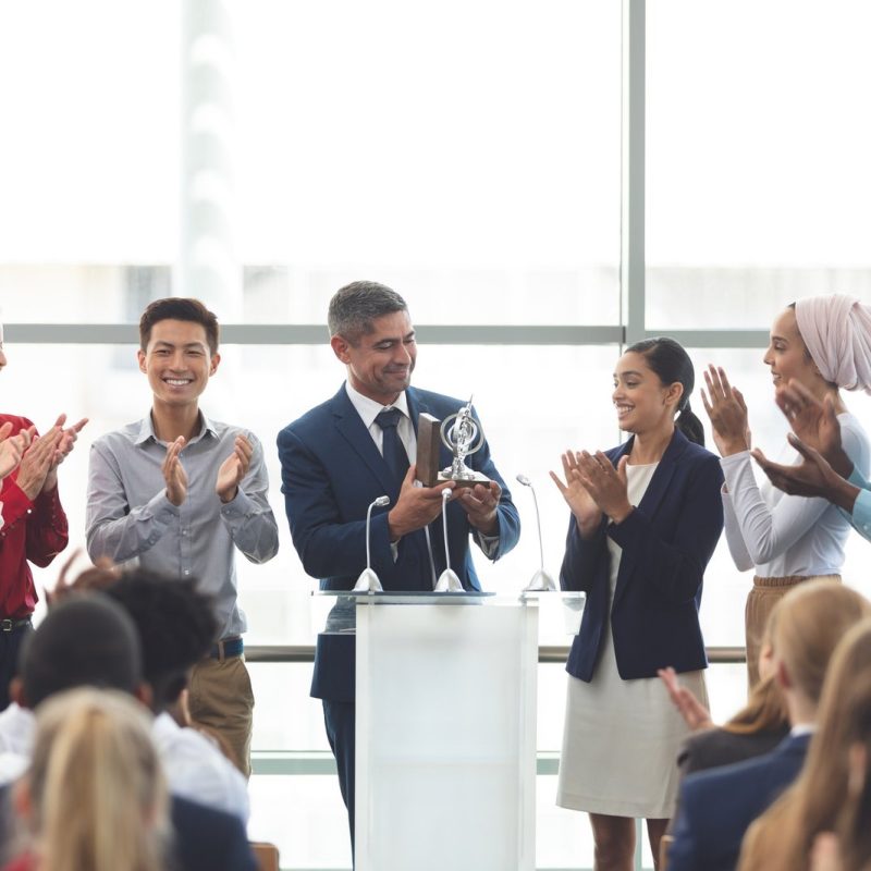 businessman-holding-award-on-podium-with-colleagues-at-business-seminar.jpg